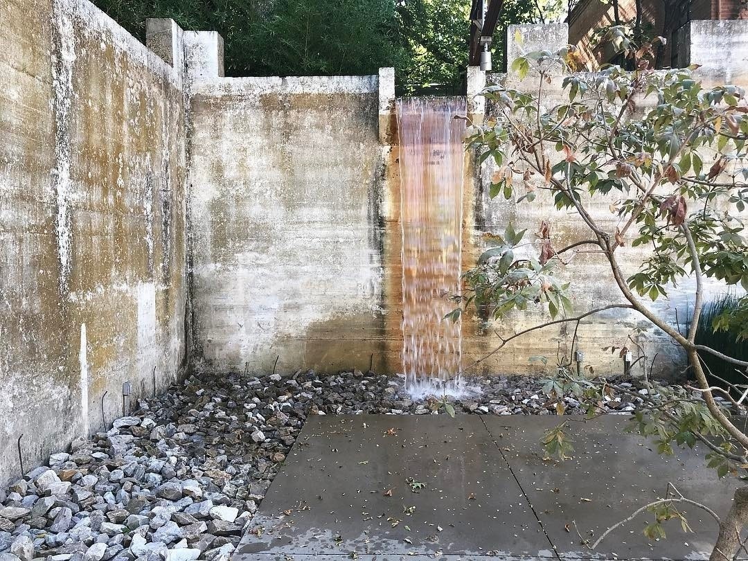 photo of Turtle Creek Water Works, view of two rundown walls with water pouring over an opening at the top of the wall straight ahead, rocks lined the perimeter of the wall which then leads to a concrete slab, a few tree limbs enter the frame from the right side