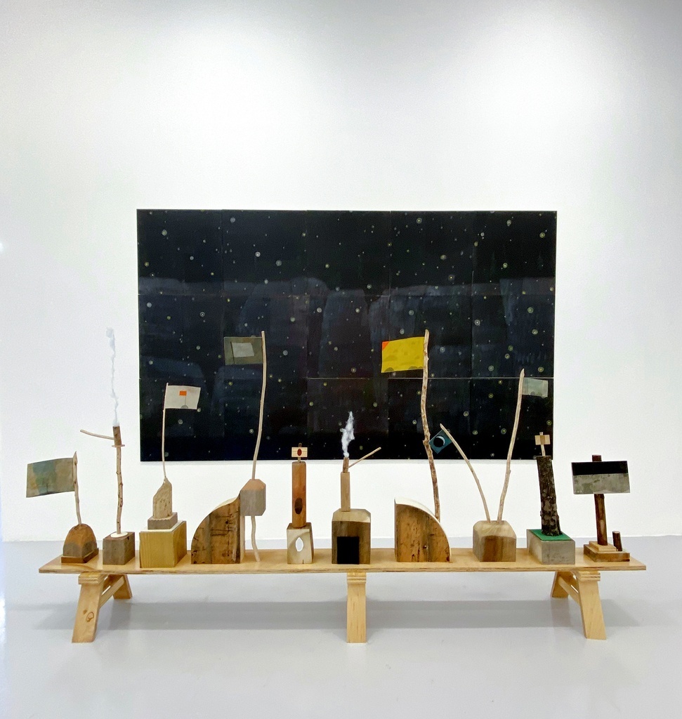A low wooden bench holds a dozen wooden totems capped by little colorful flags on tree branches. A large, dark print with a field of stars appears on the wall behind.