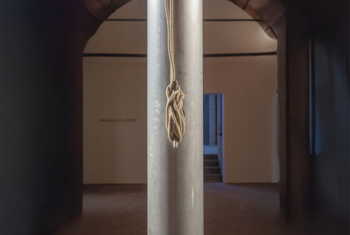 An elaborate knot on a hook on a metal flagpole centered in the frame in an arched brick-walled room
