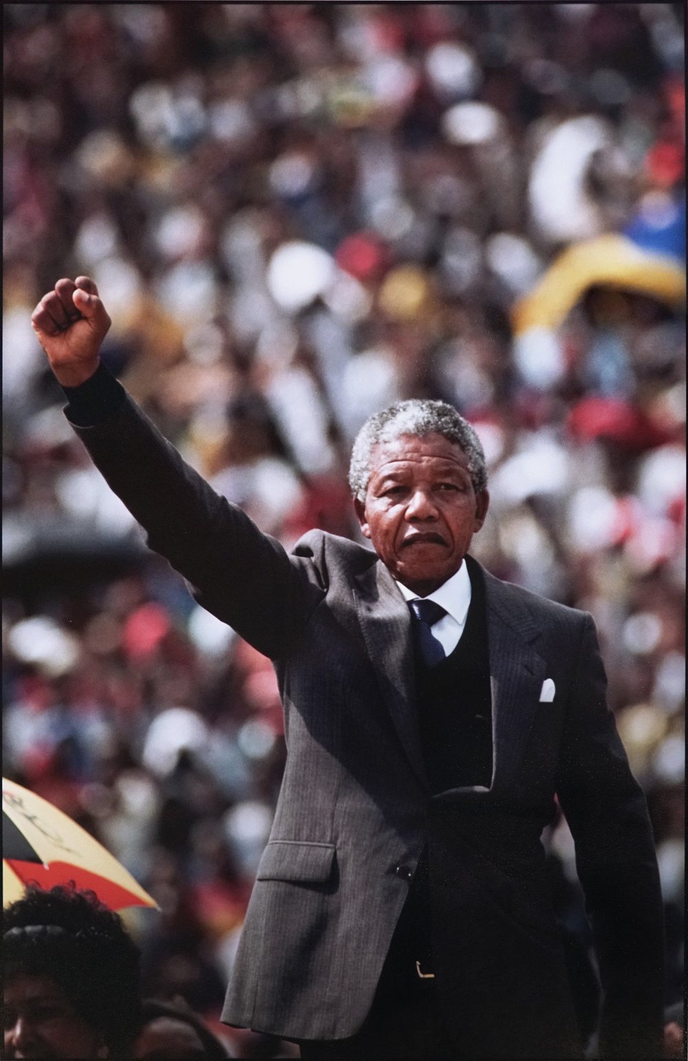 A color photograph of Nelson Mandela, a Black man with short grey hair, standing with his fist raised. Behind him, out of focus is a large crowd of people.