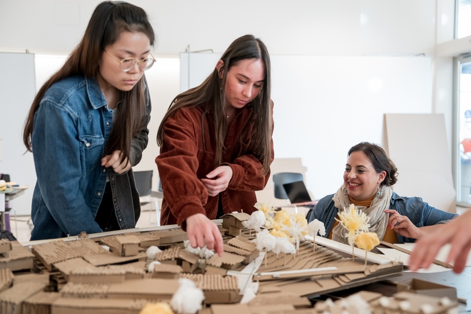 Two students speak to a smiling instructor, pointing to spots on a cardboard model of a city.