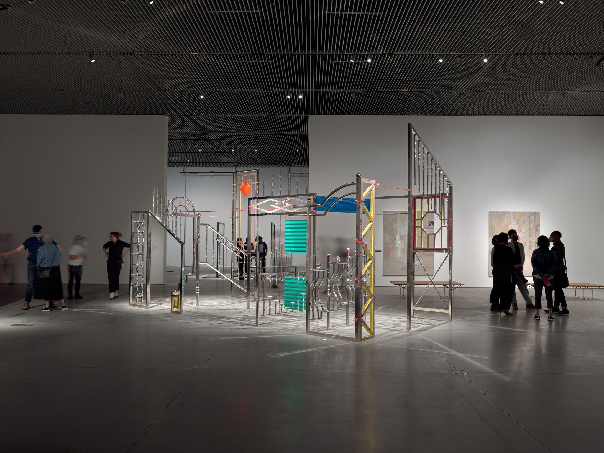 An art gallery with a large architectural installation of metal poles and railings in the middle of the room and people gathered to the side of it