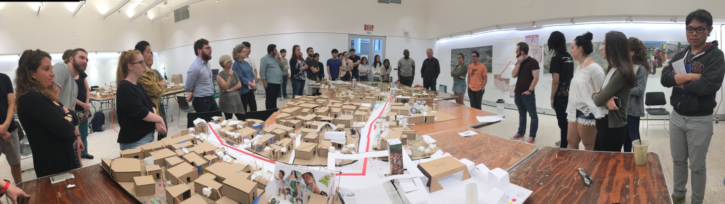 Panoramic photo of a large group of people standing around a table holding a 3D model of a city plan.
