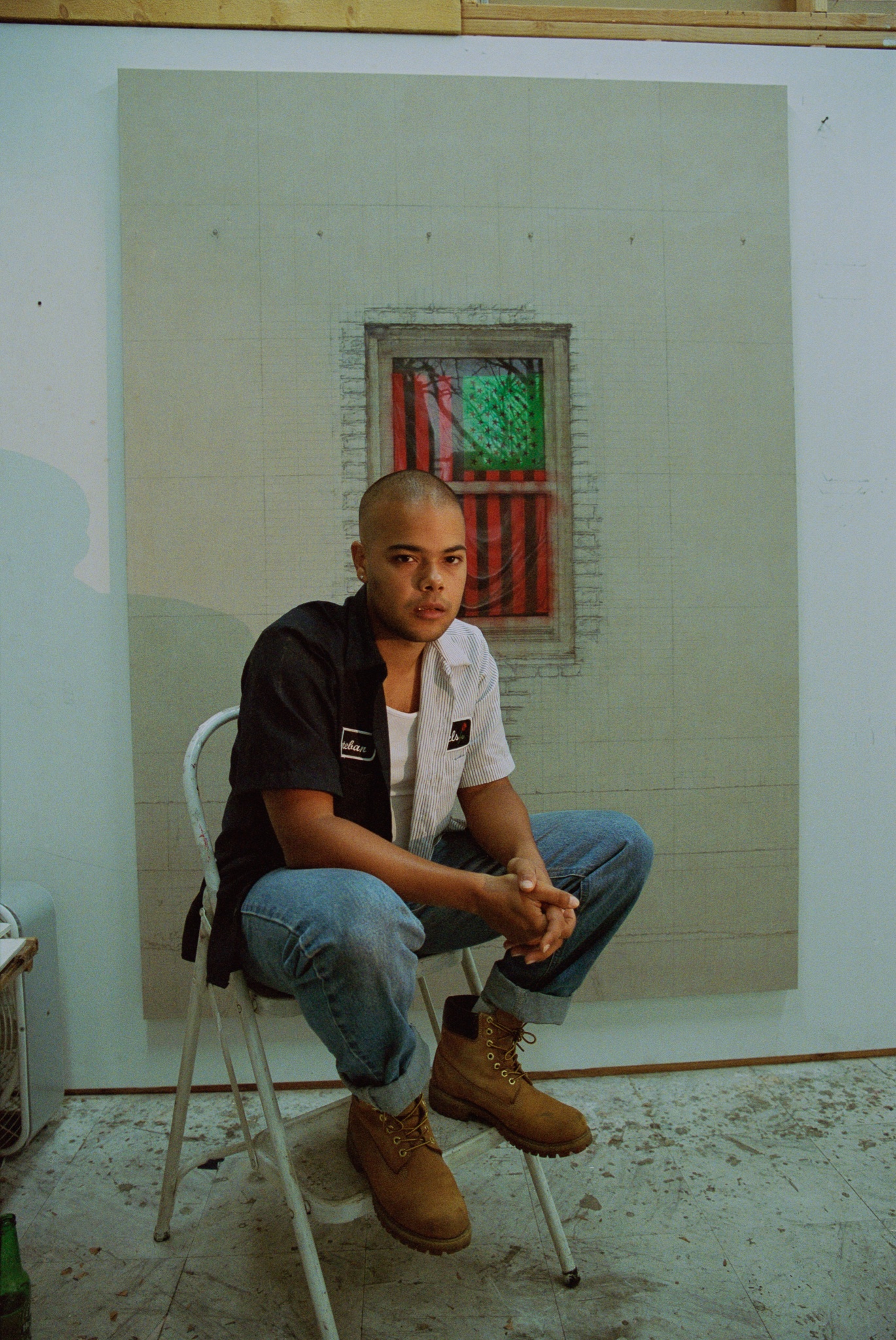 A photo of the artist Esteban Jefferson sitting in a metal folding chair with elbows on his thighs in front of an artwork on the wall. Jefferson wears jeans, boots, and a short sleeve button-down shirt over a white tank top.