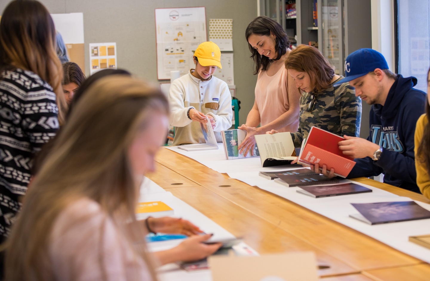 Students in a classroom leafing through design books laid out on a white sheet of paper on a wooden table. 