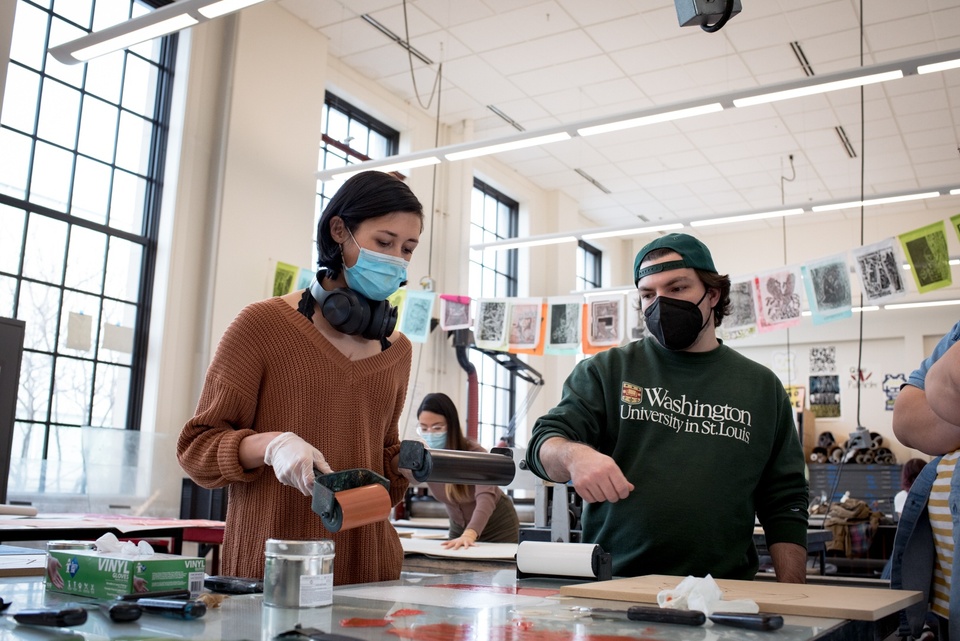 Two people stand side by side over a glass topped table. One is holding a brayer in each hand. The other is making a brayer motion with their wrist.