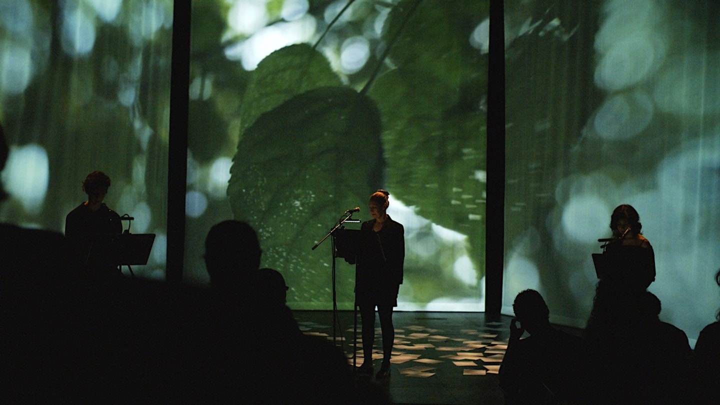 A live performance by Olynyk titled "A Cure for Immortality," featuring a performer lit at a microphone in front of a large video projection of green leaves, flanked by silhouetted performers and viewed by an audience.