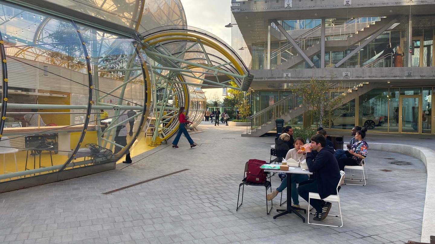 Exterior photo of people outside at a grey, tiled plaza; a contemporary, round-walled building with glass windows is to the left.