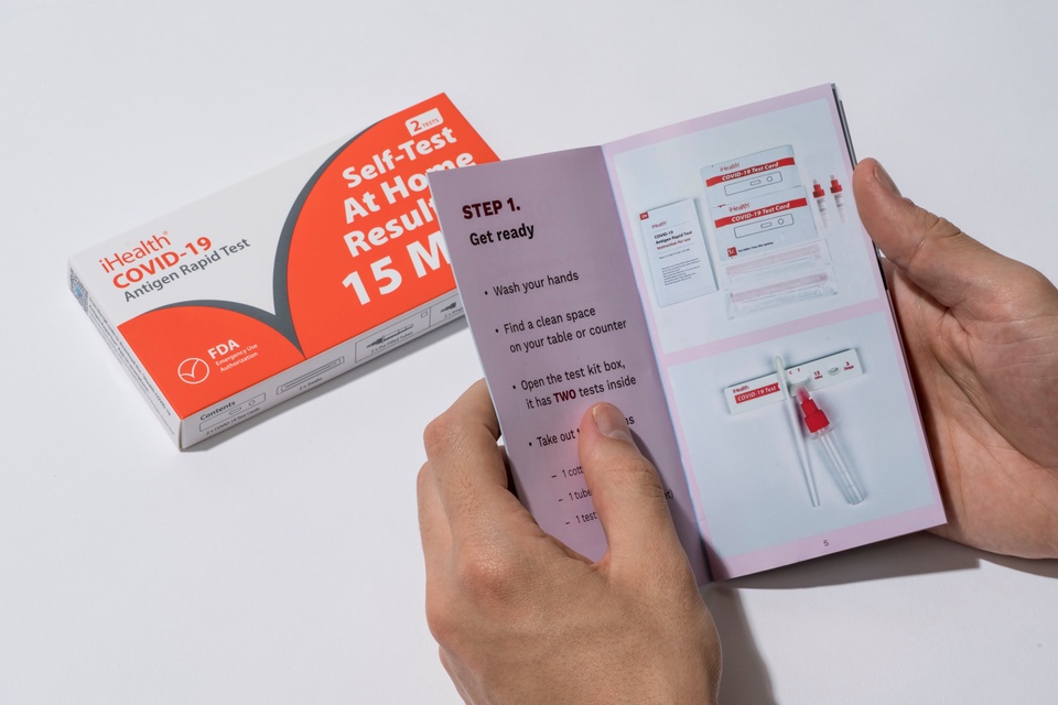 Two hands hold open the booklet to Step 1 of using an iHealth self-testing kit. An iHeath kit is on the table.