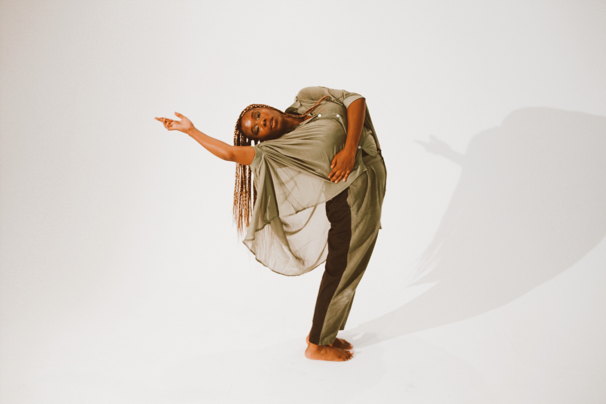 A Black person with long braids bends over backward with one hand outstretched away from their head and body. They wear a draped, sheer fatigue green blouse and pose in what appears to be a photographer's studio with a blank, neutral backdrop.