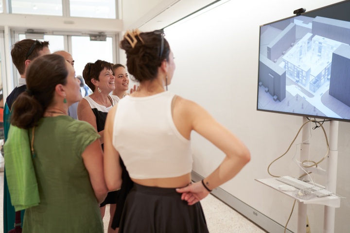 A group of people view an architectural rendering on a tv screen. The rendering features a low, square glass-walled building set in a courtyard surrounded by other buildings.