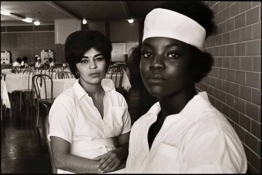 A black and white photograph of two women staring directly at the camera in matching uniforms seated in a cafeteria-like setting.