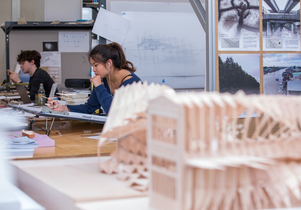 Close-up of basswood model. In the background two people work at spaced out drafting tables.