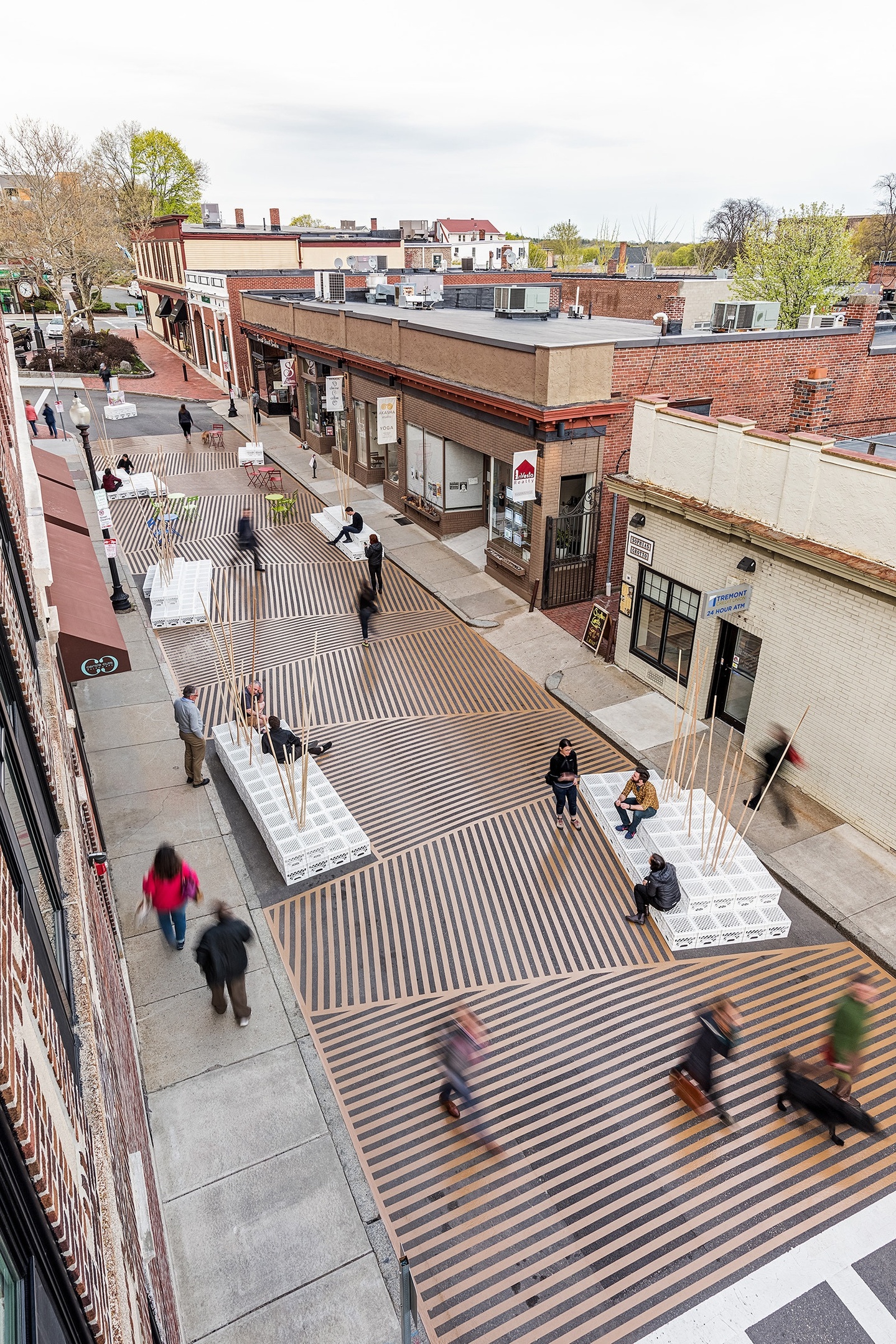 Overhead view of people walking on a pedestrian throroughfare with shops/buildings on both sides. Between the sidewalks is a walkway made of interesting black-and-tan striped boards set up in different patterns, along with block-like white-and-gray furniture pieces to sit on.
