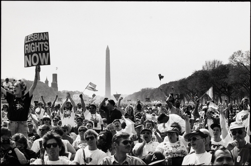 A black and white photograph of a large obelisk in the distance behind an expansive crowd waving flags and signs with the most prominent reading “LESBIAN RIGHTS NOW.”