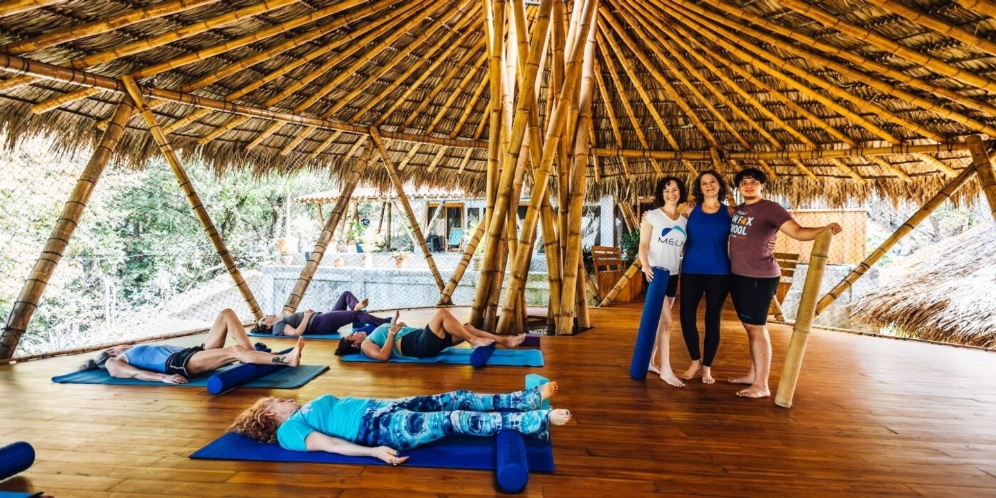 4 women laying on yoga mats and 3 people standing looking at the camera inside of a hut