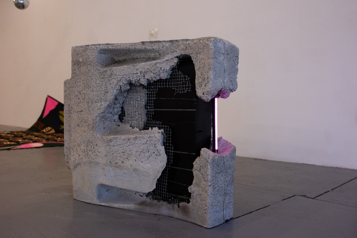 A roughed-up flatscreen tv is half-encased in wire mesh and concrete, sitting on its side on the floor. A neon pink lighting strip runs along the outer edge of the tv screen where it is exposed.