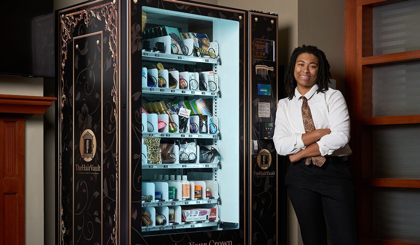 A person leans against a vending machine of hair products and smiles at the camera with their arms crossed.