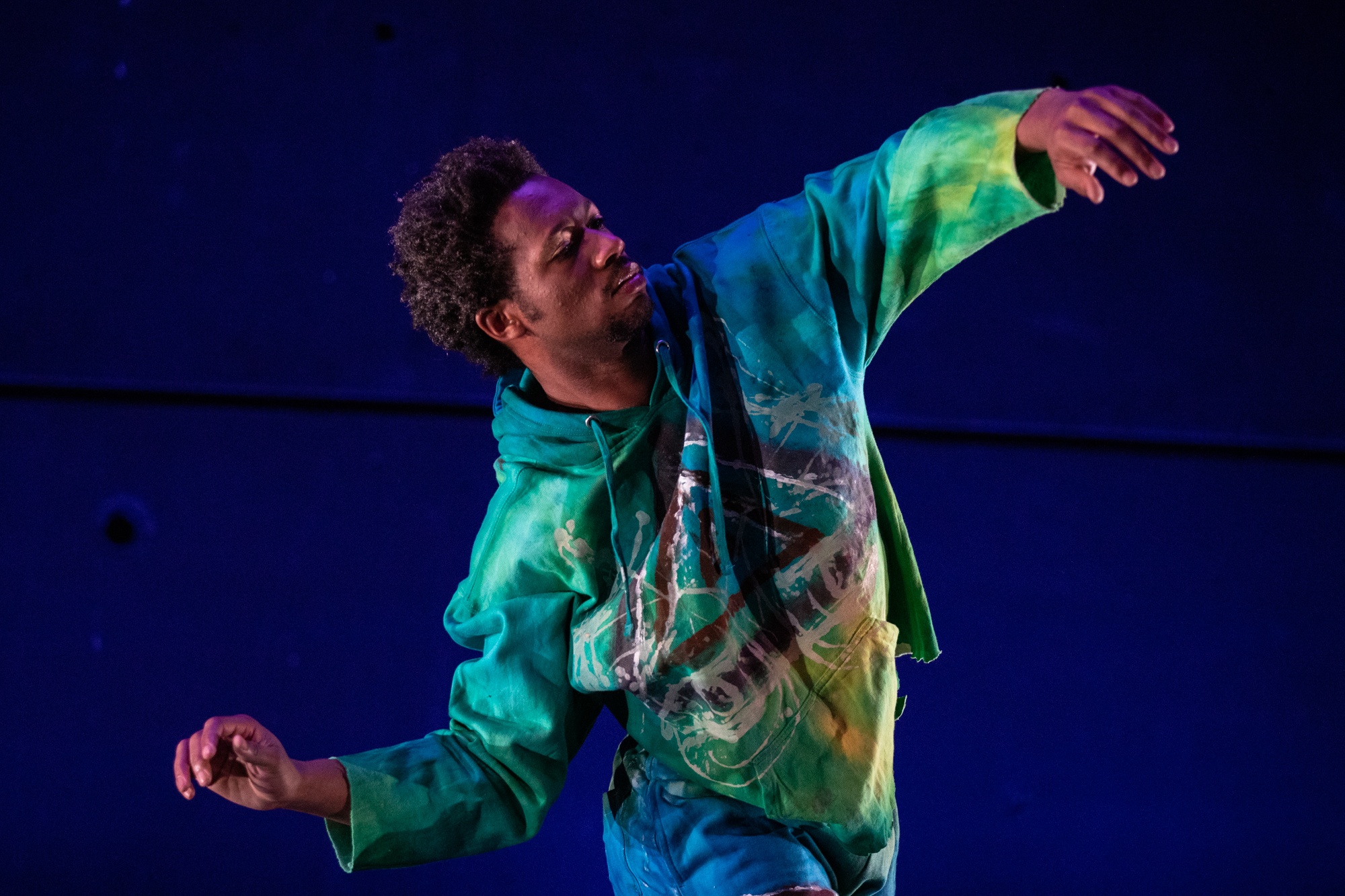 A Black male dancer in a green, blue, and yellow tie-dyed sweatshirt leans slightly backward with arms outstretched and his left arm raised and right arm lowered to the level of his waist