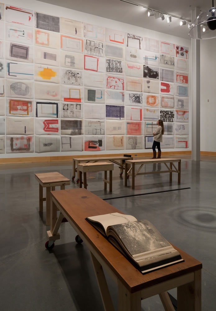 Printed works tiled, landscape orientation, on a tall white wall: some compositions are rendered in warm, pink/coral/peach/orange hues, while others are in black and white. Five wooden tables/platforms positioned in the center of the space, the closest one to us displaying a book, which is open on a spread containing a grayscale photograph.