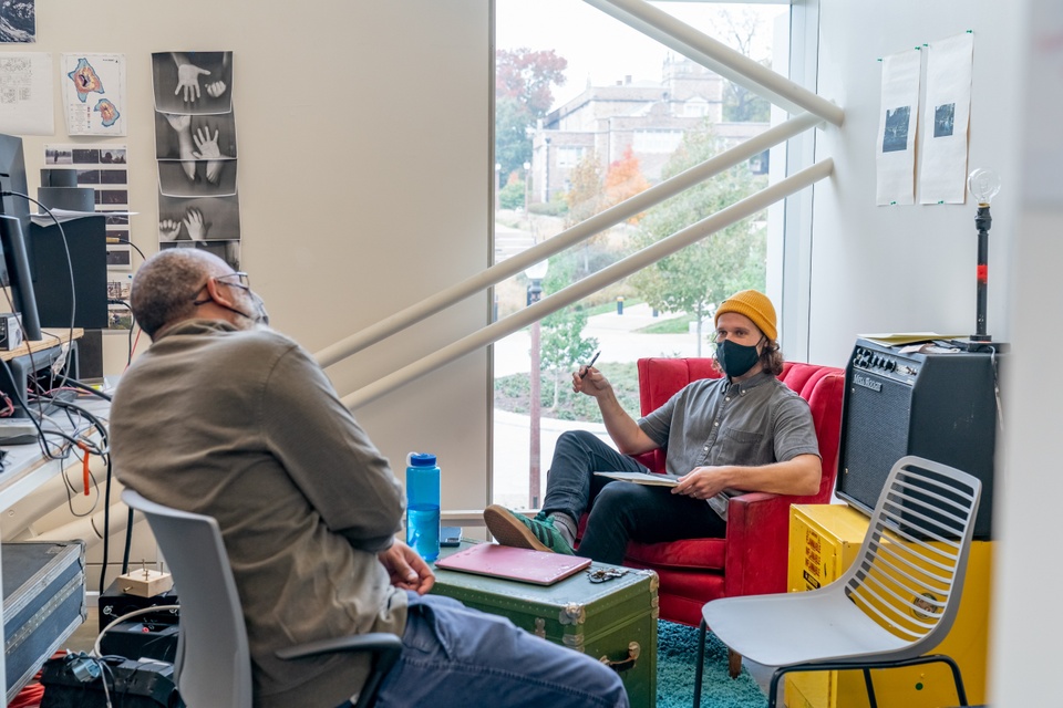 Two people chat in a studio space with a tall window and exposed beams. The studio is arranged with a teal shag carpet, a red armchair, yellow hazardous materials cabinet, and artwork pinned to the walls.