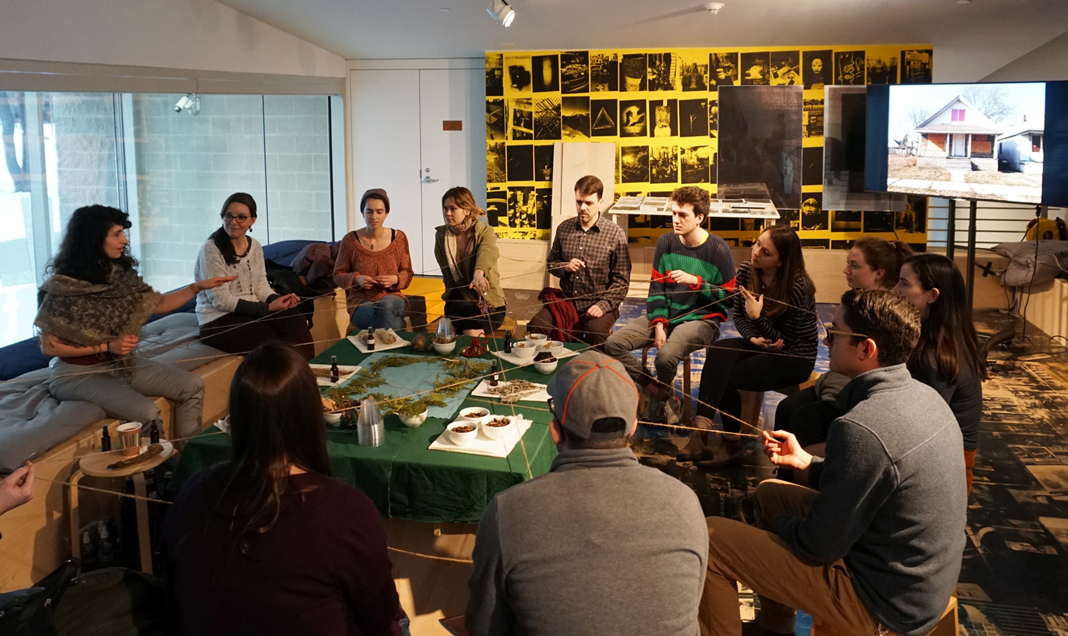 A group of people sit around a table covered with plant and earth materials holding a string that connects them all.