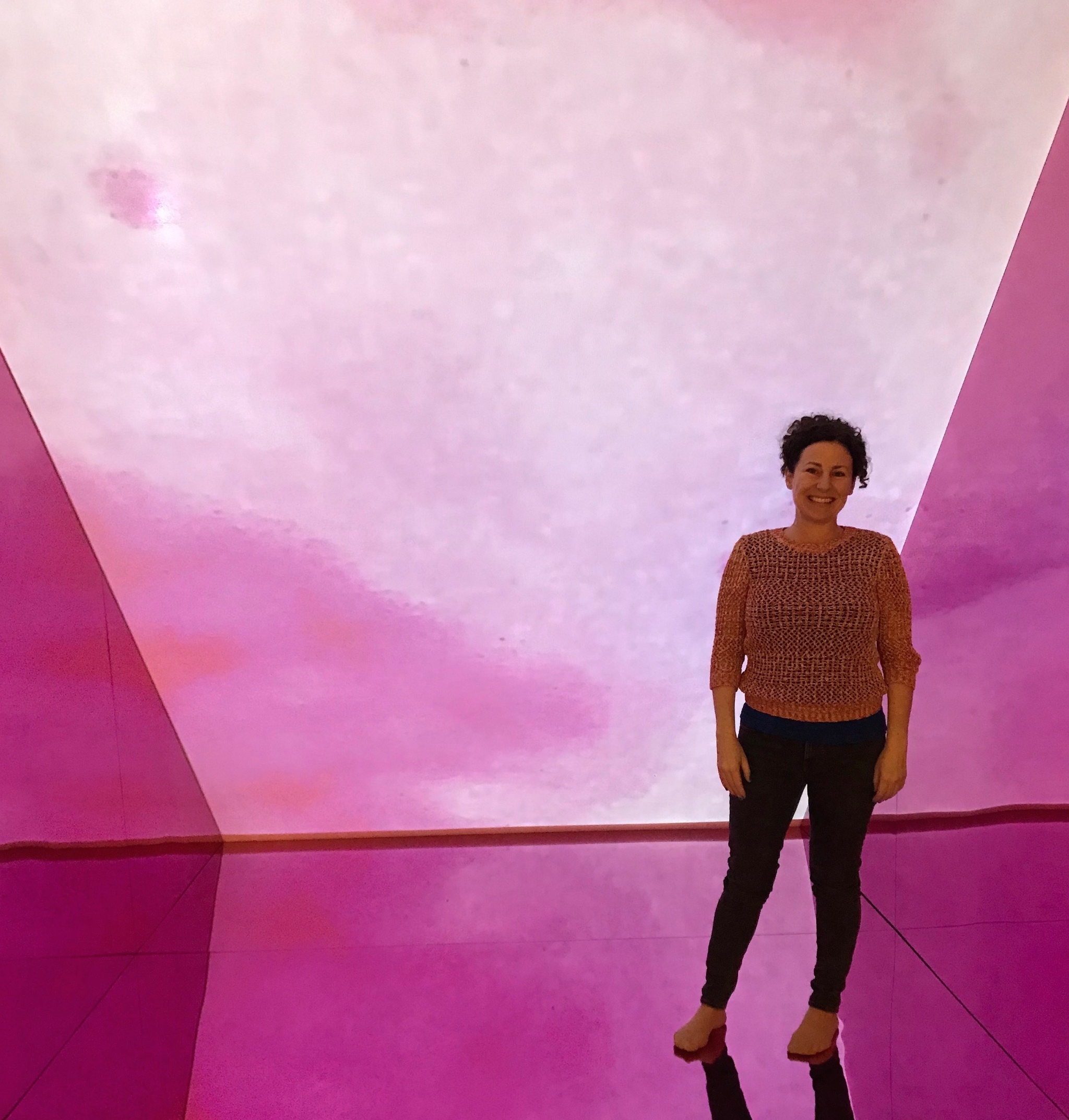 A photo of Mara Mills standing in an art installation of pink projections on the walls and floor. She stands facing us and smiling. 