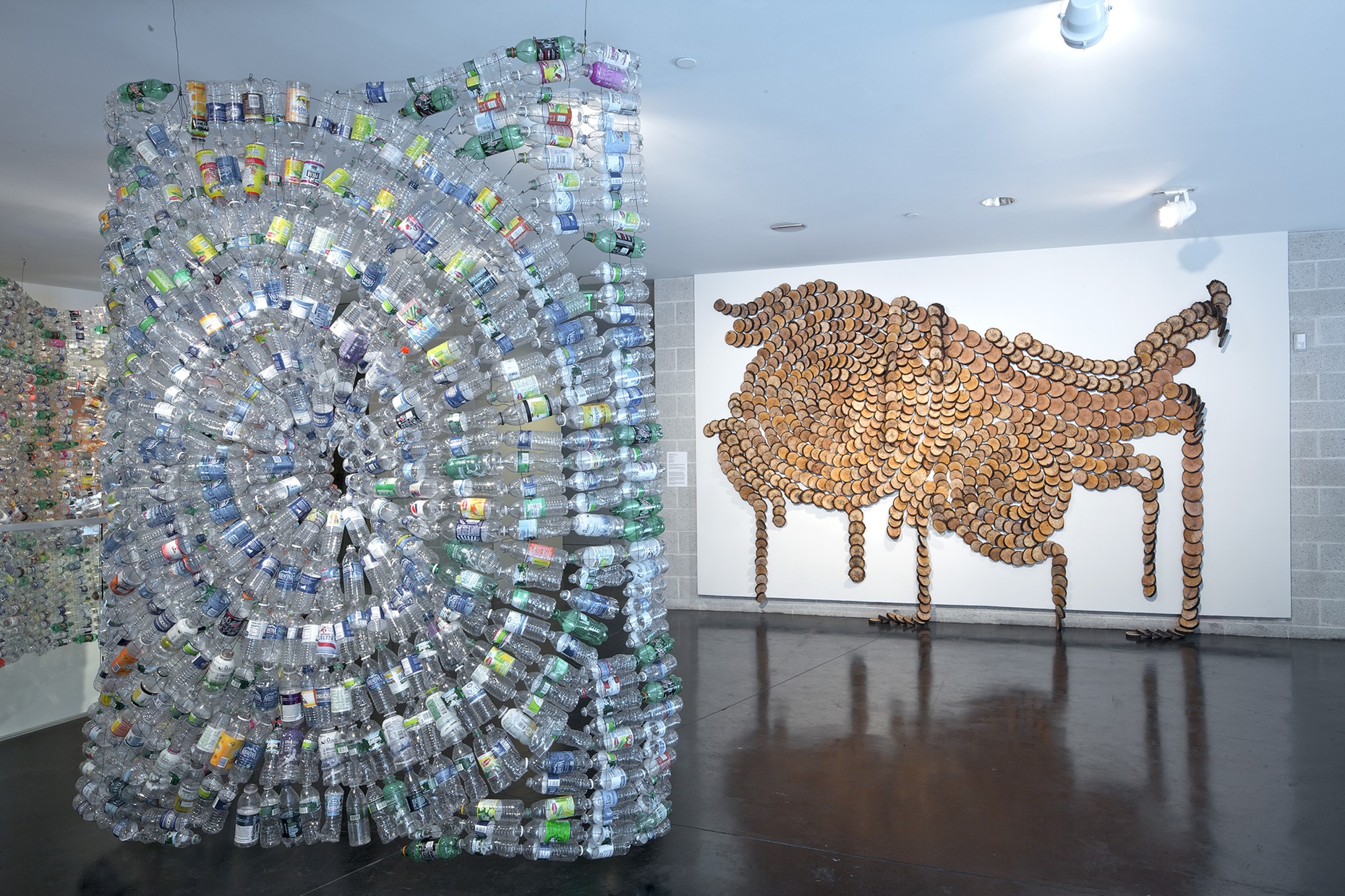 A swirly, three-dimensional sculpture made of plastic bottles in front of a white wall with an abstract design made of wooden circles.