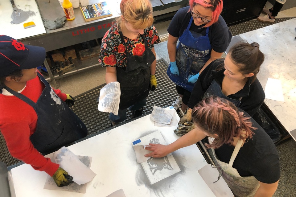 Four printmaking students wearing aprons work on Duane Slick’s helper print on a large table.