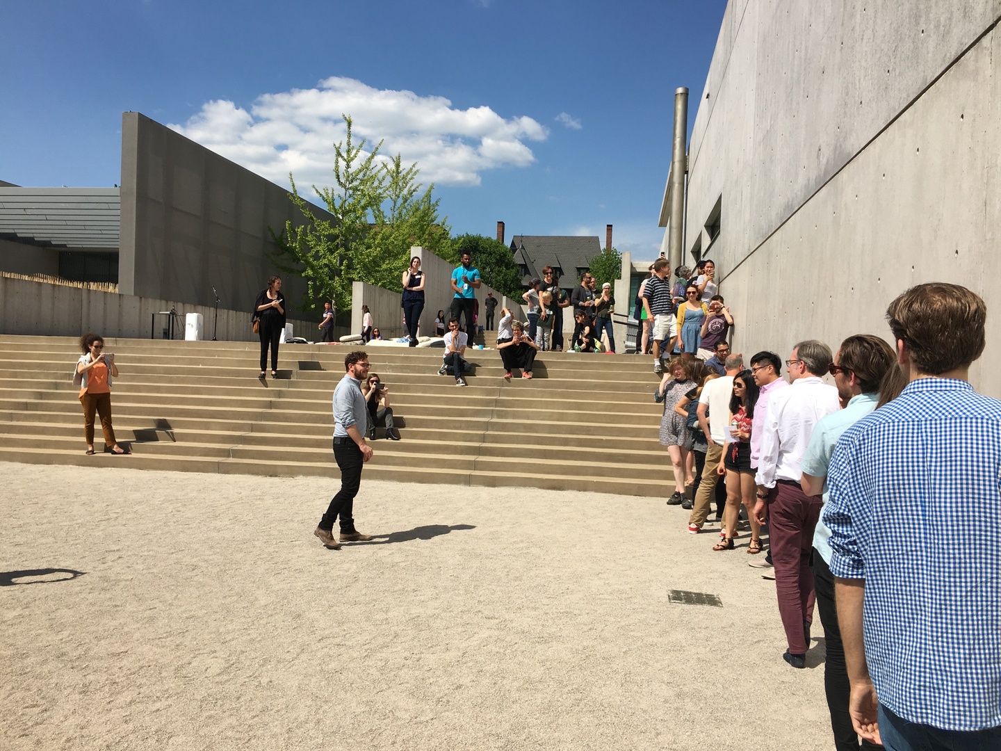 Group of people in a graveled courtyard lined up along a concrete wall.