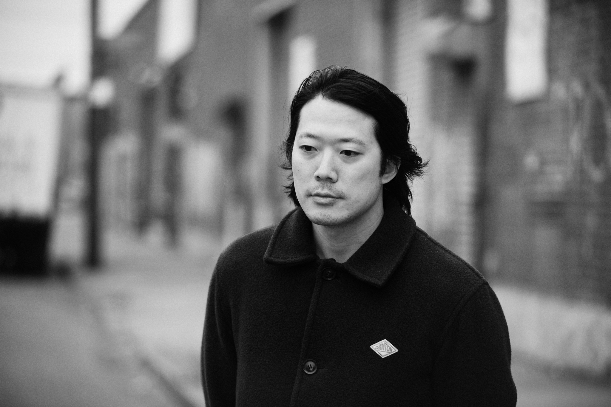 A photo of the artist Kenneth Tam on a street with blurry warehouse buildings behind him. Tam wears a dark wool coat and has his hair tucked behind his ears.
