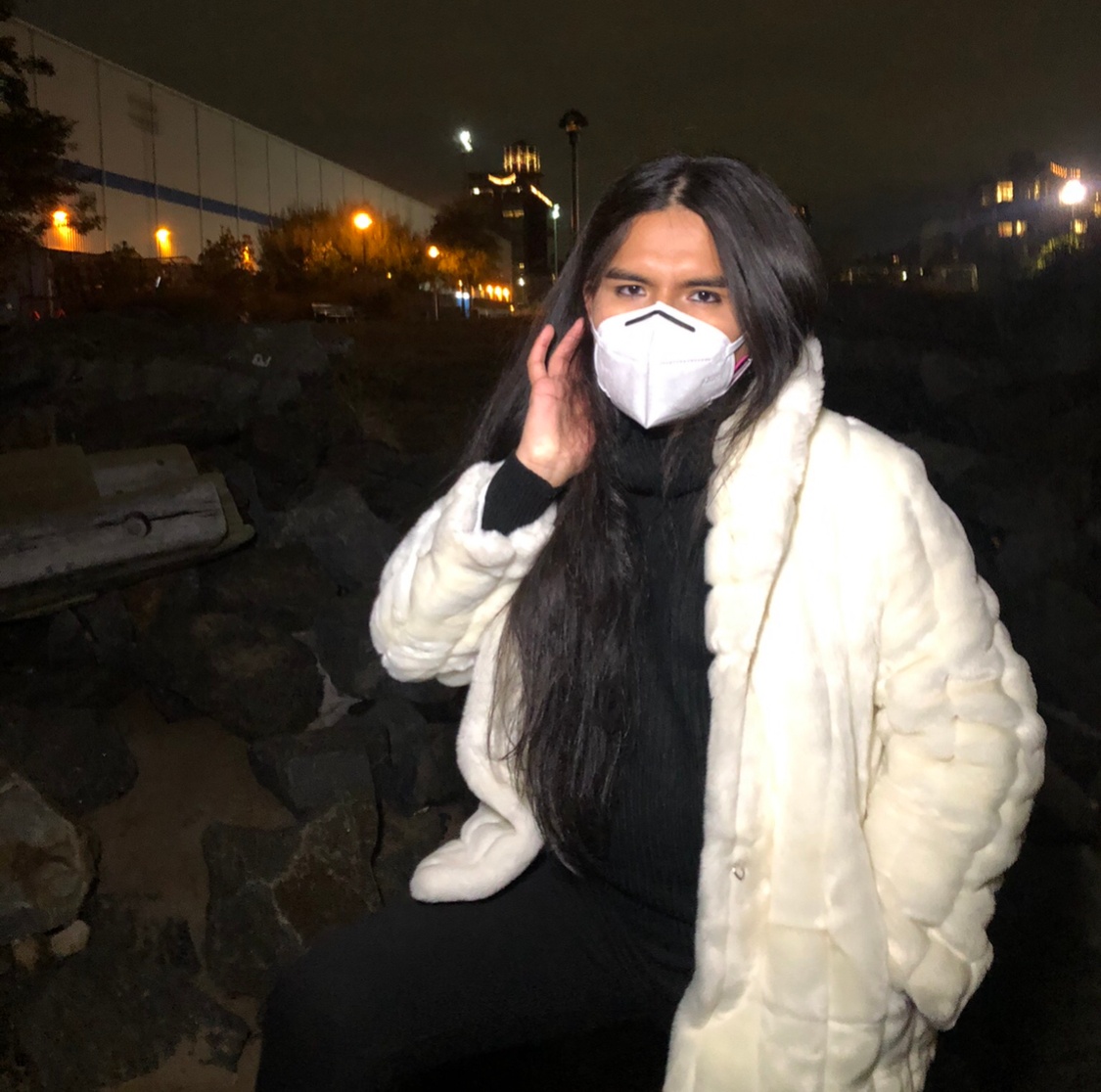Poet Jimena Lucero stands outside at night wearing a white face mask and plush coat. Lucero holds one hand behind her right ear with her long hair falling over her shoulder behind it. 