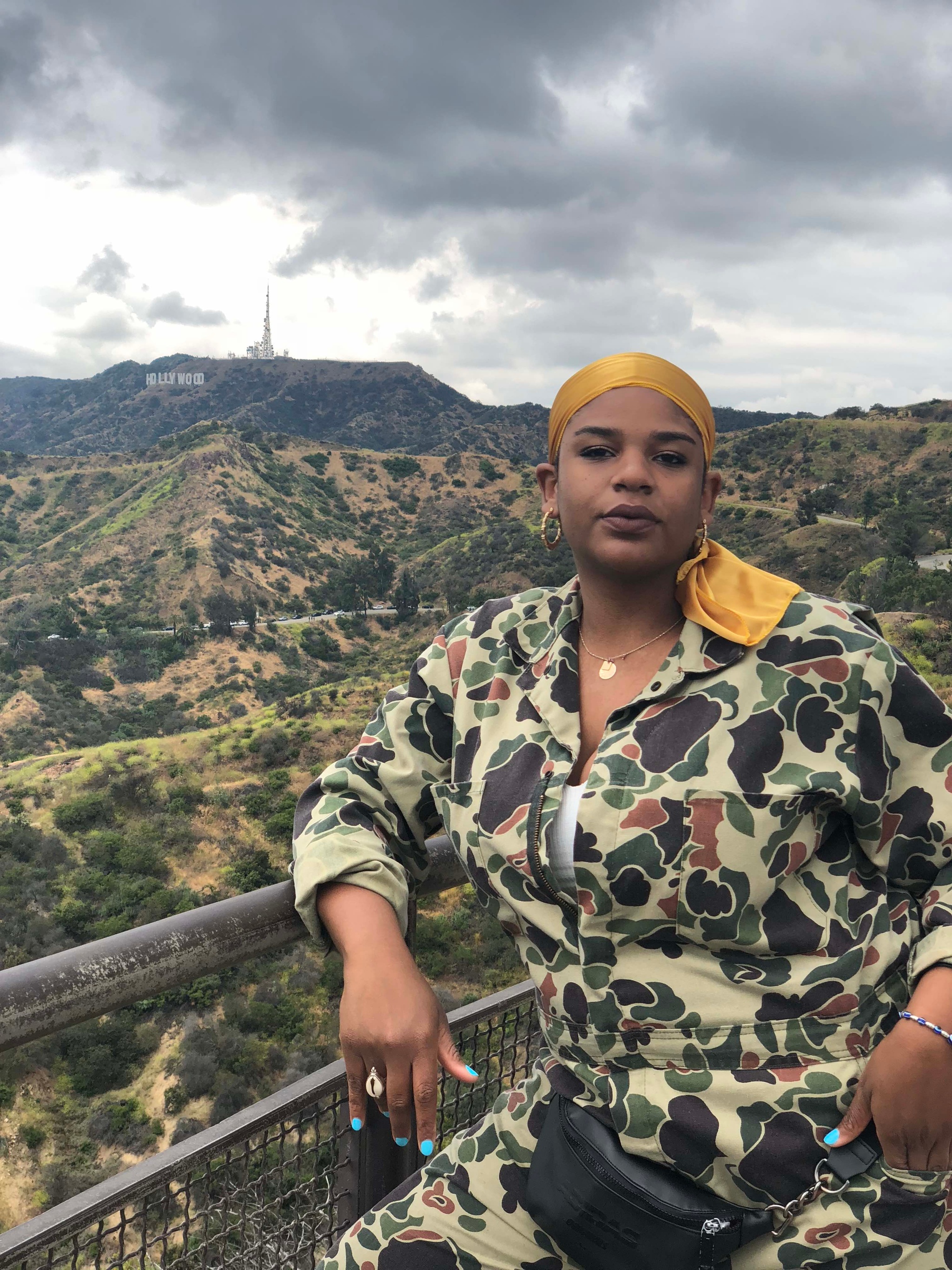 A person dressed in a camo outfit that blends into the hilly landscape behind them. They wear a yellow scarf around their head and lean against a railing with one elbow propped against it. 