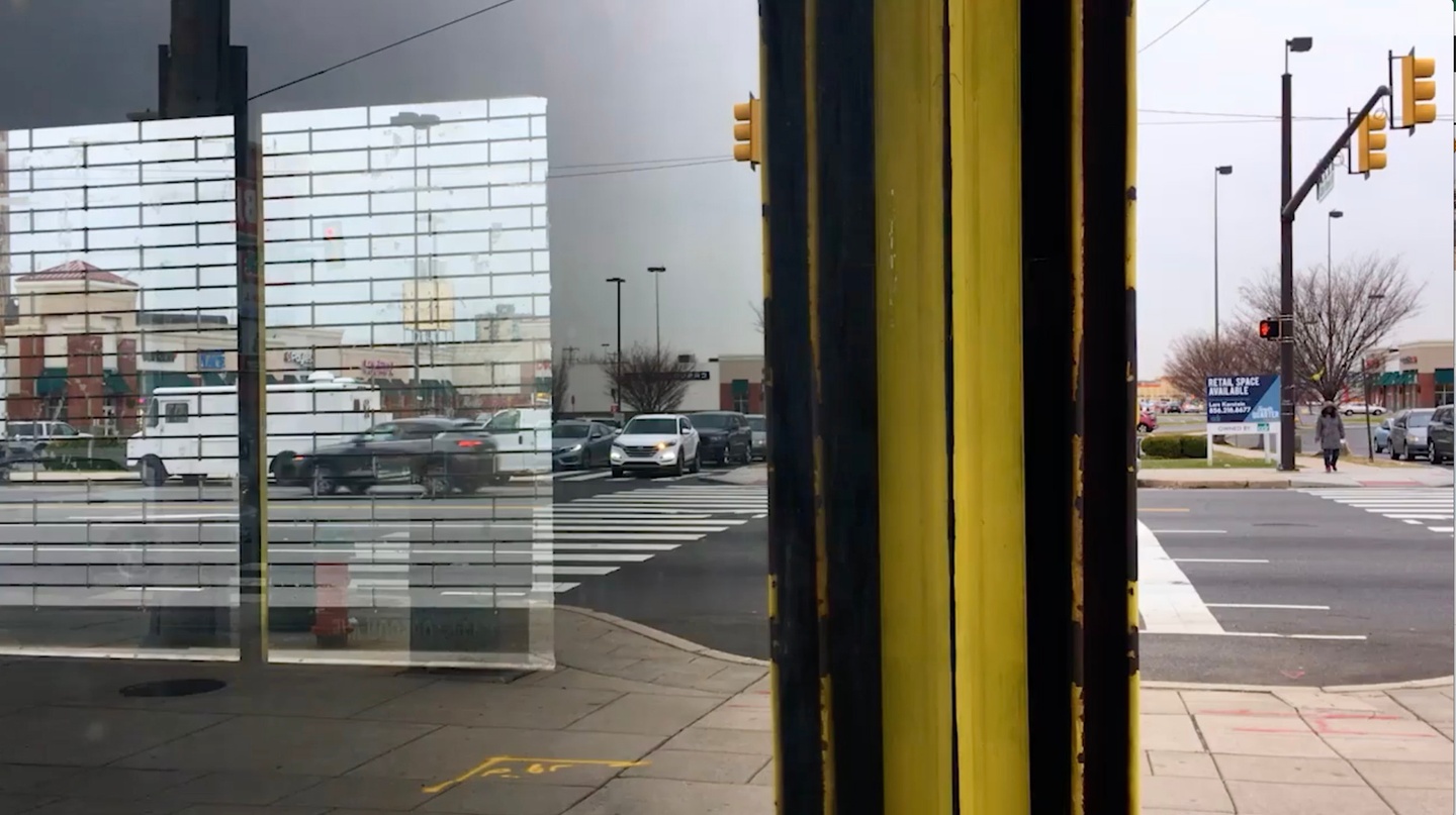 Video still, looking out at people walking on the street; a yellow and black pole interrupts the frame near the middle, while a transparent brick overlay is visible on the lefthand side.