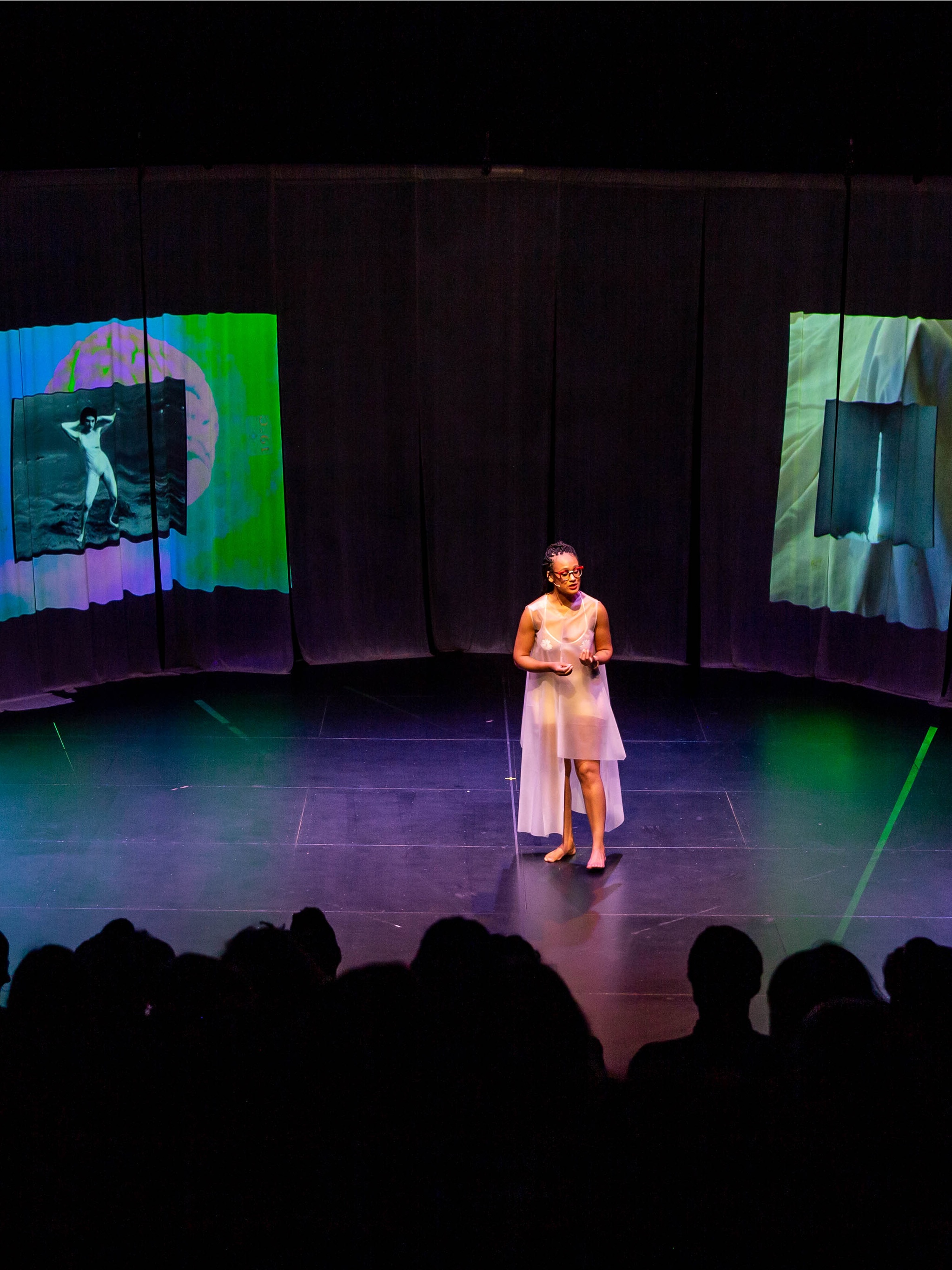 The artist Eleanor Kipping performs on stage in a darkened theater in front of an audience. She wears a sheer pink dress and red framed glasses. She is speaking to the audience, flanked by two screens with video projections.