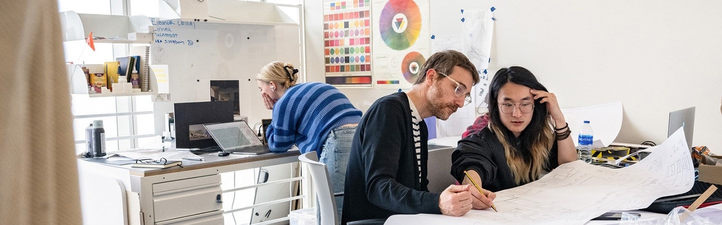 Two people at a desk looking at architectural drawings in a studio space. Another person is behind them working at a laptop.