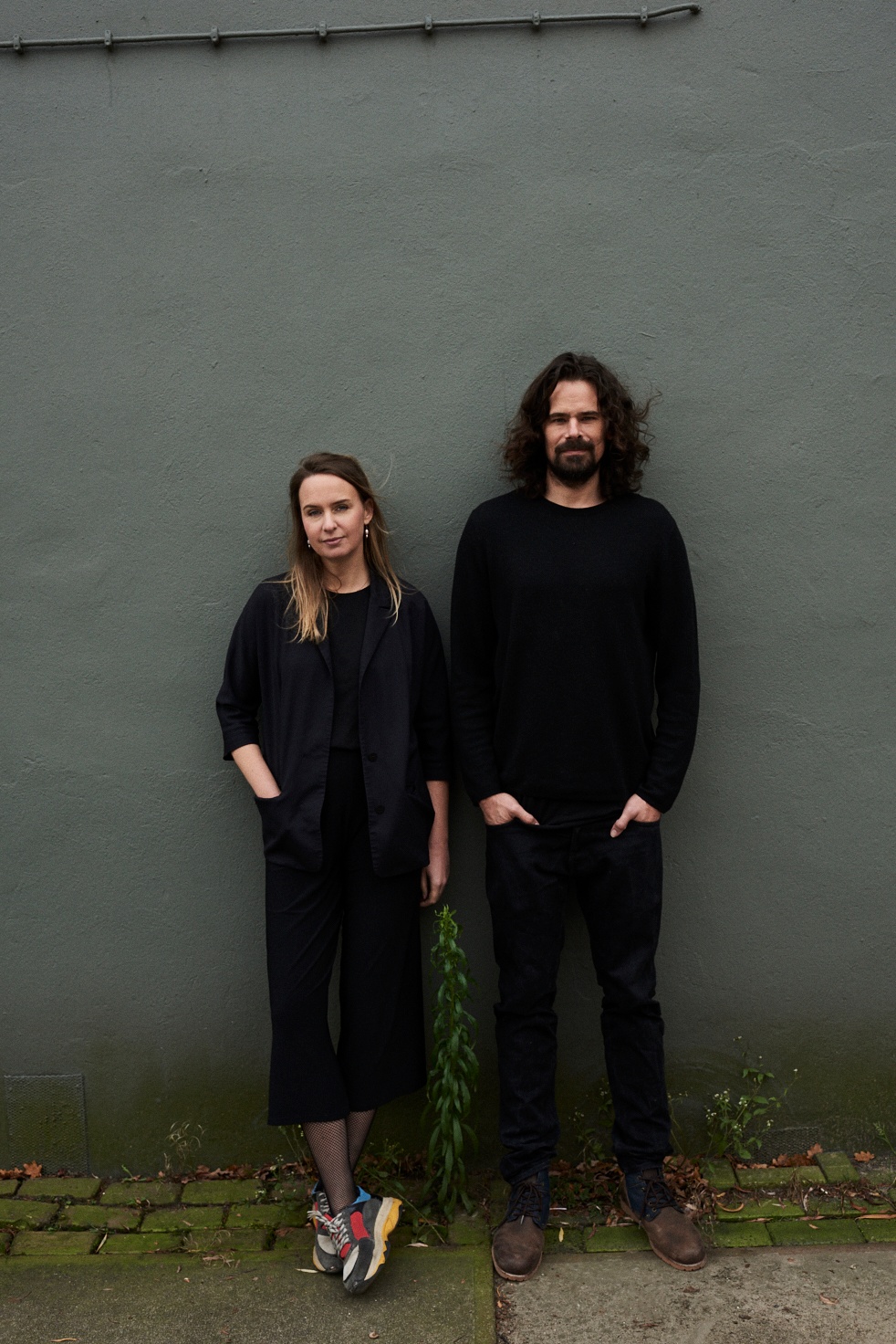 The artists of DRIFT, Lonneke Gordijn and Ralph Nauta, standing in dark outfits side by side against a dark gray wall.