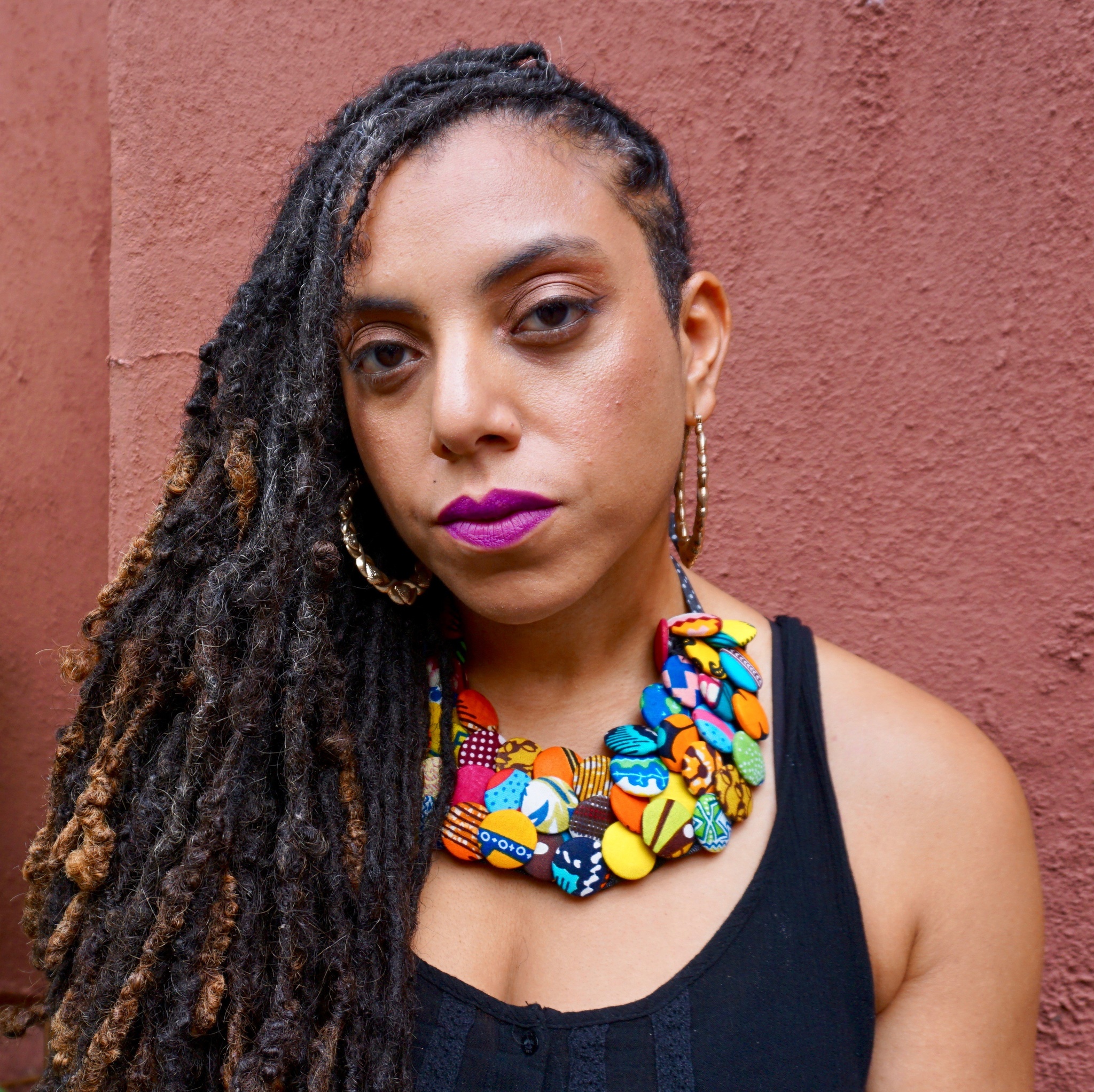Artist Nia O. Witherspoon poses against a burnt-red wall. She looks into the camera while wearing a wide multicolored necklace made of overlapping discs and has her hair falling over the right side of her face and shoulder. 