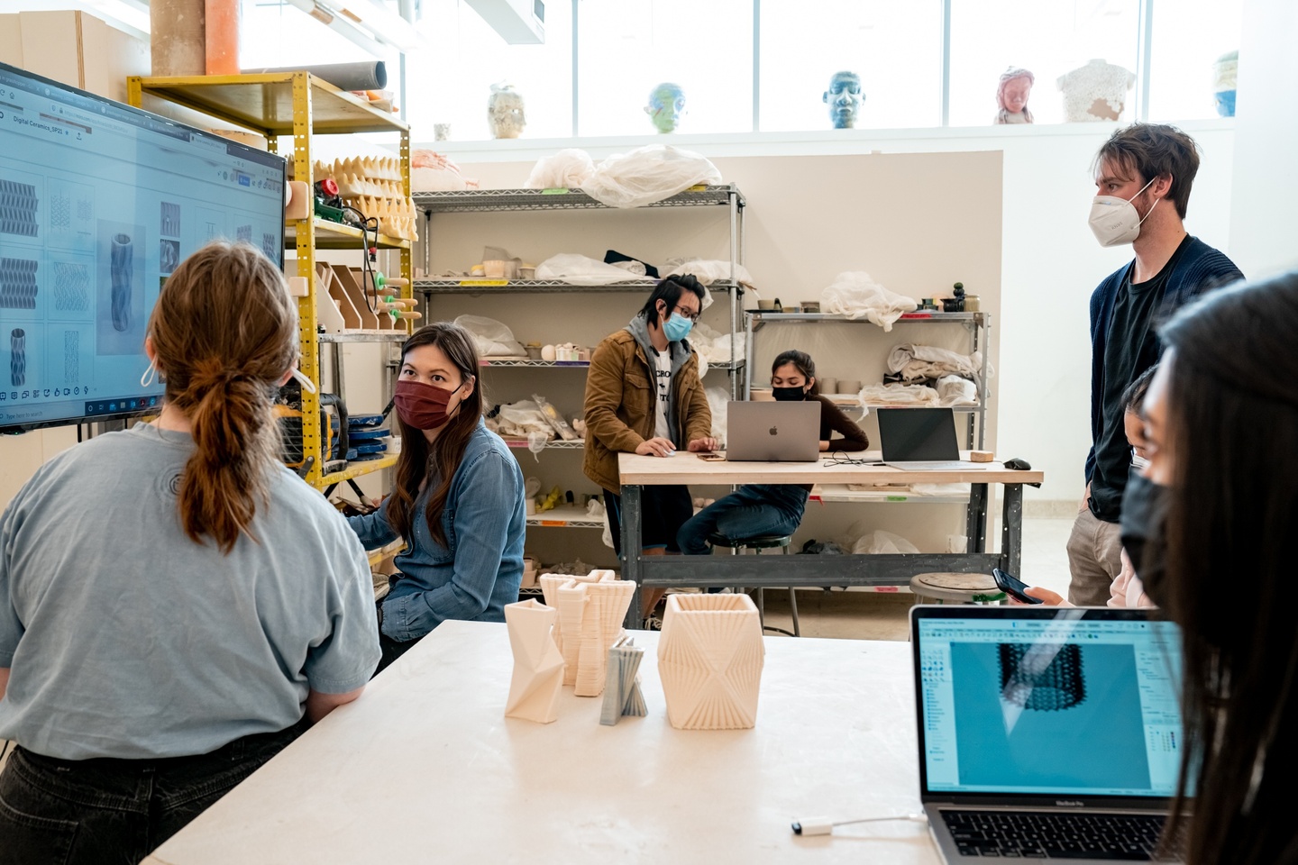 Person instructs several people on 3D printing using a large monitor. Three 3D-printed ceramic objects sit on the table next to them. The back wall has shelves lined with clay and other ceramics tools.
