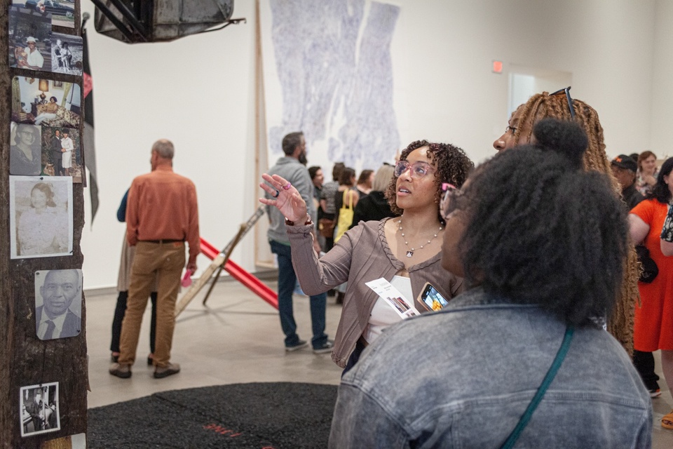 People in a gallery space look at a railroad tie with vintage photographs of a family nailed to it.