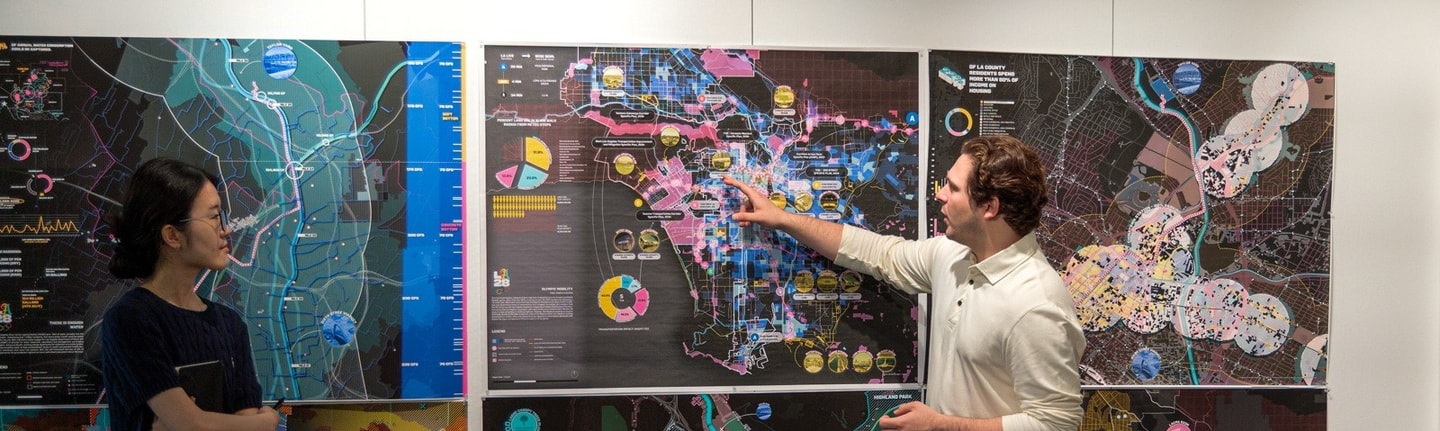 Two students stand to present their work during a pin-up. One watches while the other points at the map on the wall.