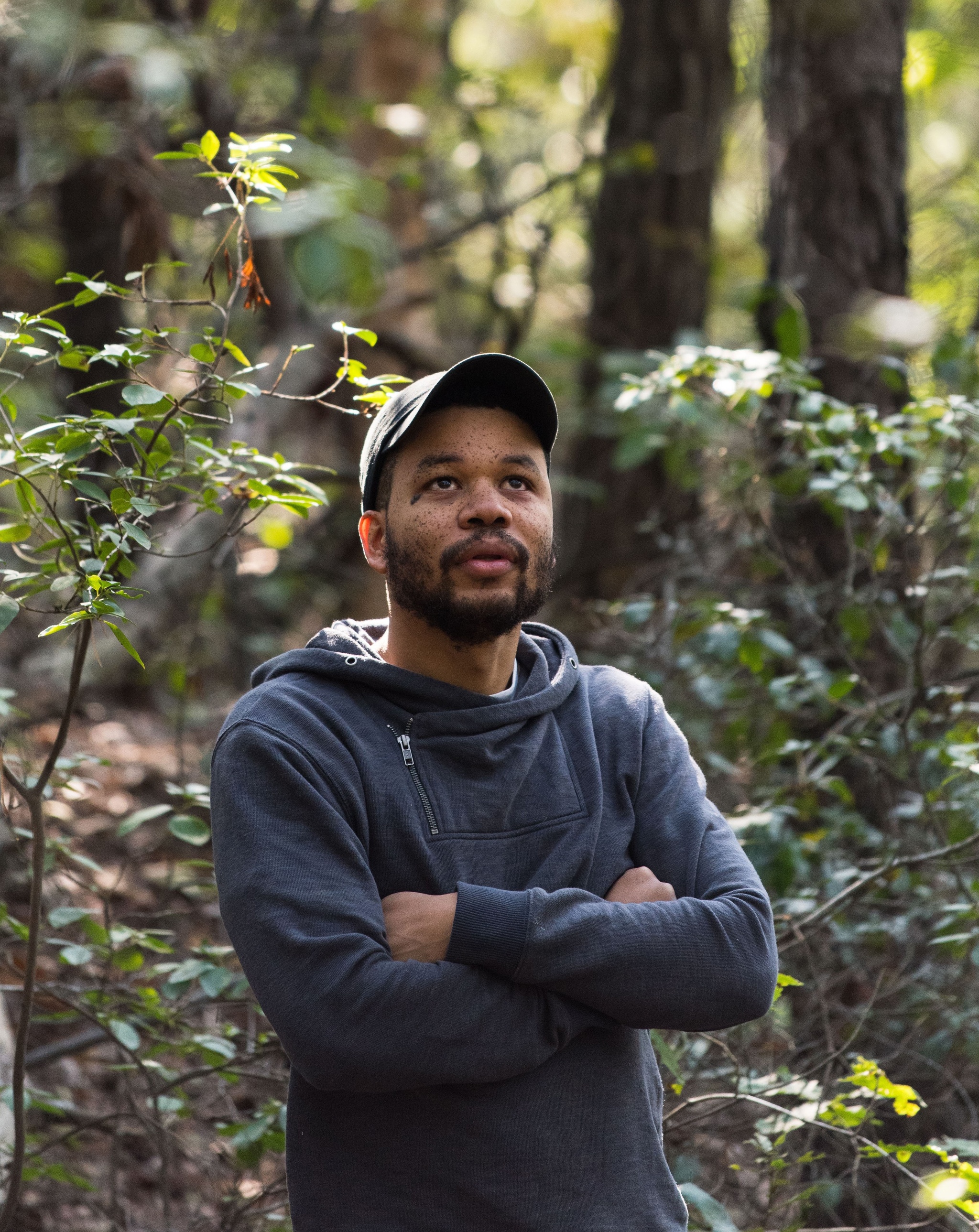 A portrait of Collision/Coalition artist Oscar Murillo in a forest looking up at trees