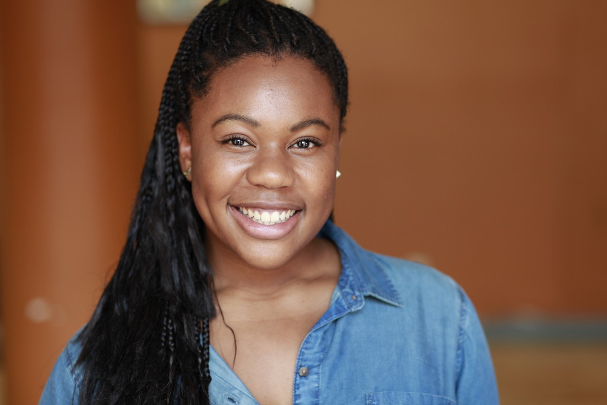A Black woman with long hair swept over her right shoulder smiling and wearing a denim button down shirt against a blurry brown-orange wall in the background