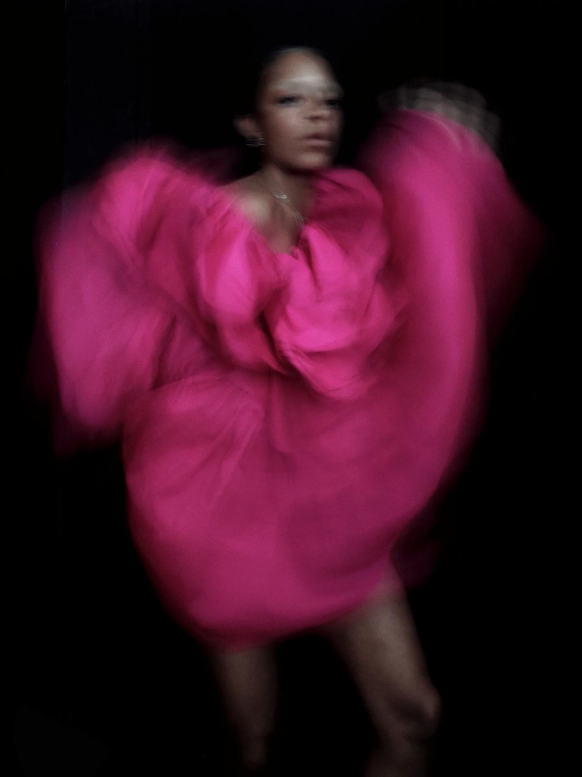 A blurry portrait of artist Kelsey Lu against a dark background. They wear what appears to be a pink dress that is fanning up and out with their movement. 