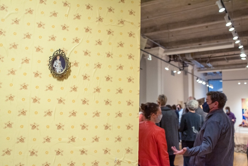 On the left half of the frame is a yellow wallpapered wall hung with a tiny oval portrait of a person in armor. Around the corner of the wall is a gallery space filled with people.