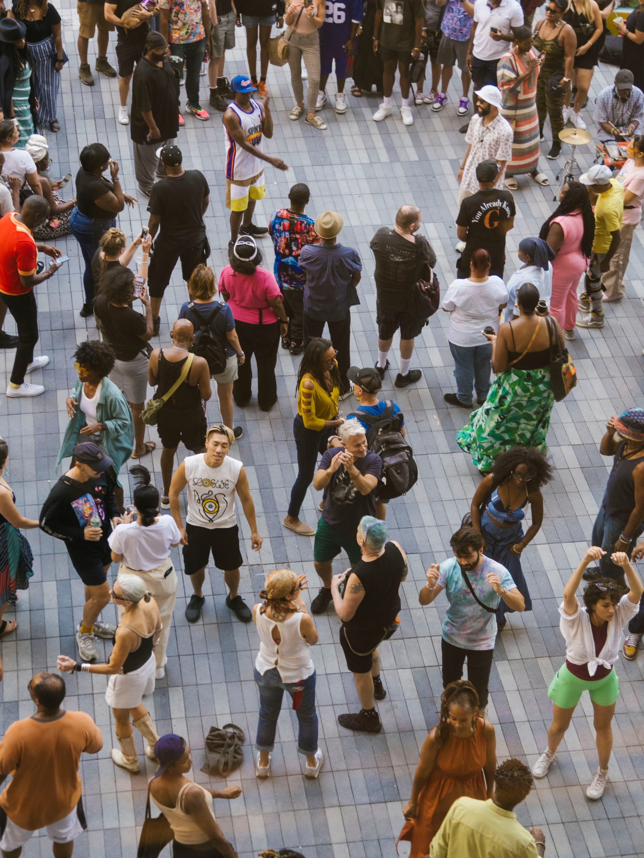 A crowd is seen from above on an outdoor patio with gray paving stones. People are dressed in colorful tshirts and are dancing. One audience member looks up at us. 