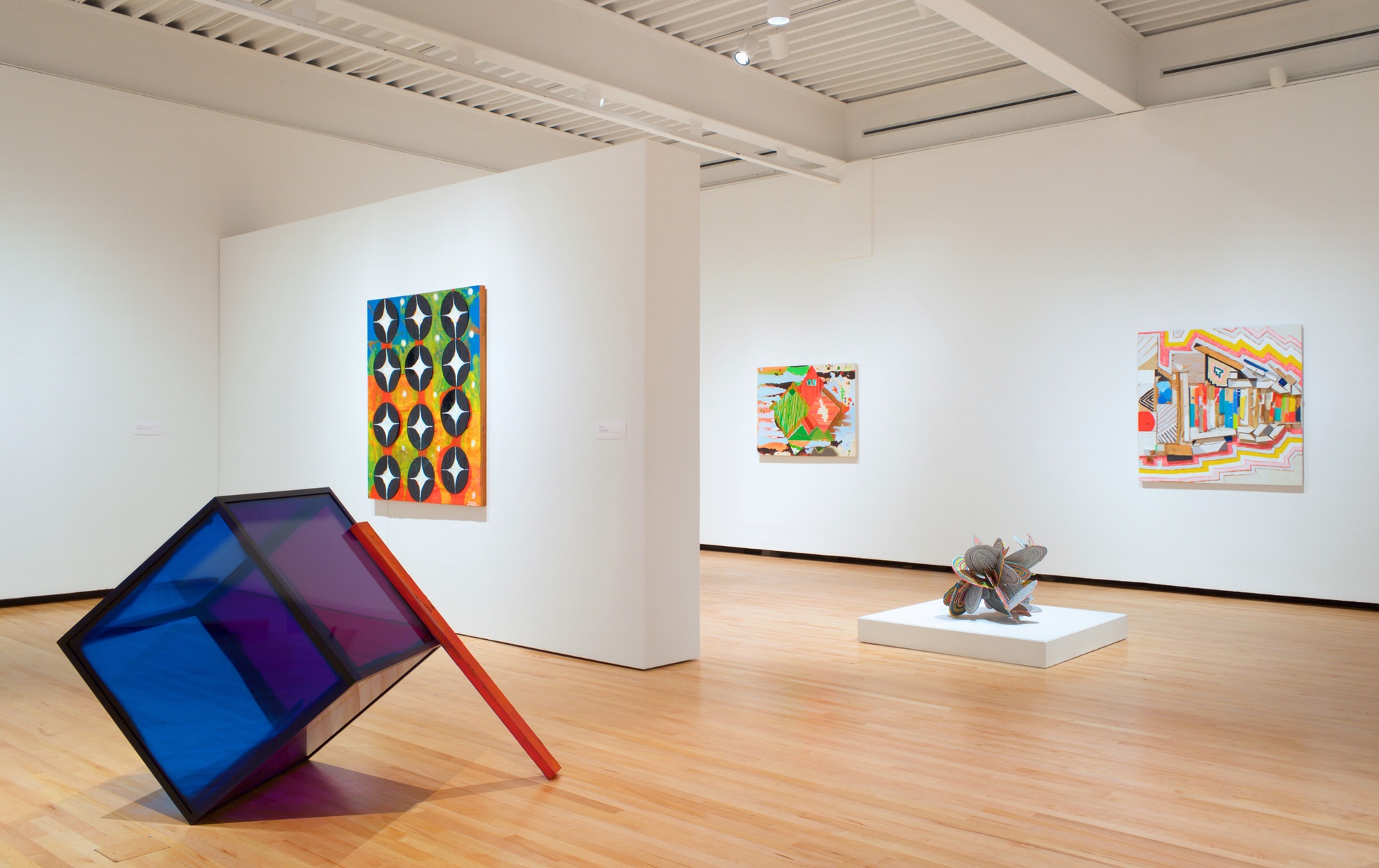Various abstract art in an illuminated room and to the left is a purple and blue cube tilted at an angle with a red stick leaning against it.