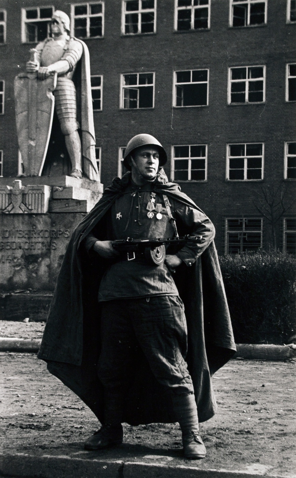 Black and white photograph of a man wearing military uniform holding a gun and posing with his hand on his hip, with a sculpture of a man in a suit of armor in the background.