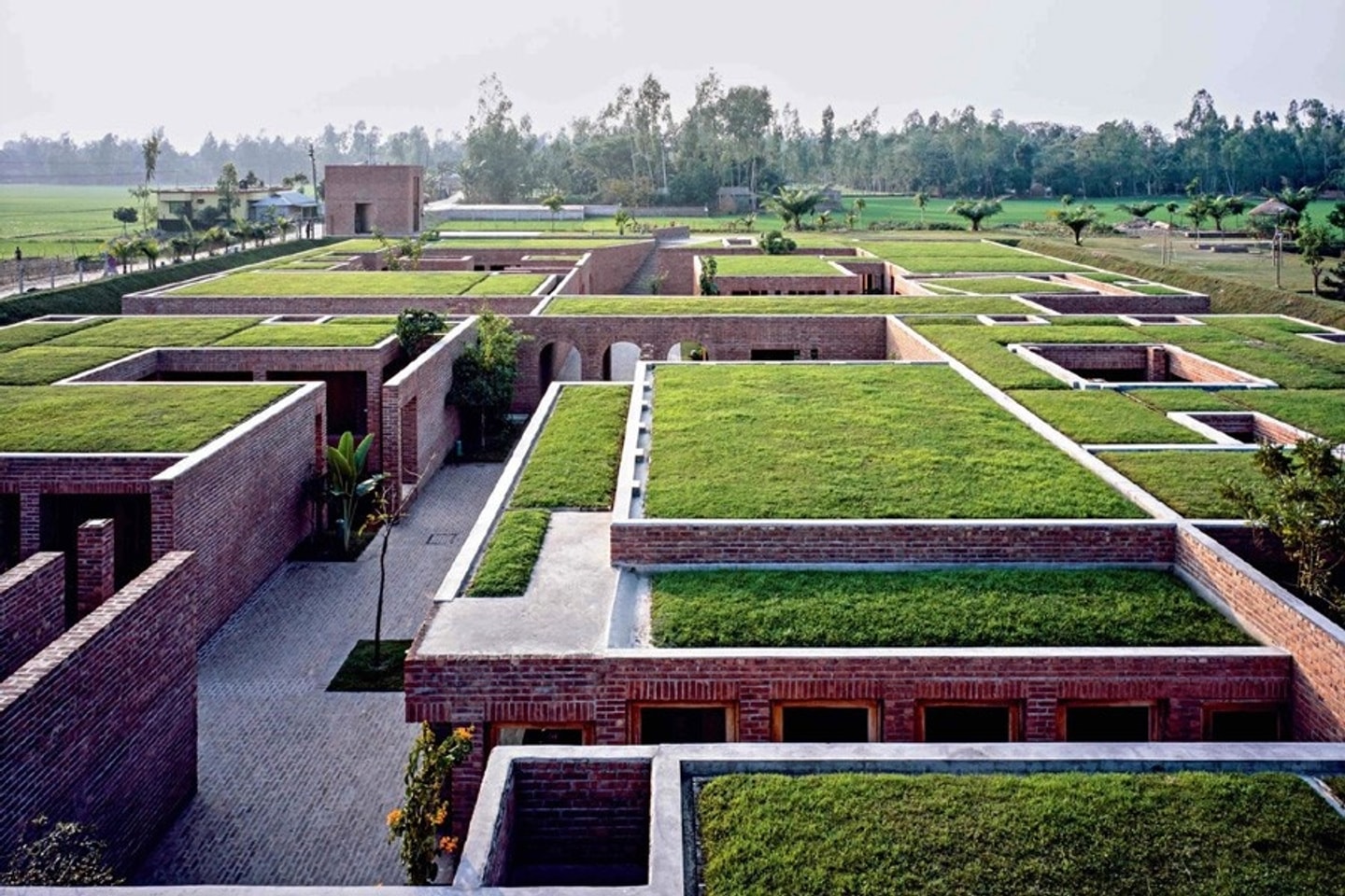 An elevated view of a low-rise red brick architectural complex featuring green roofs covered in grass, open courtyards, and brick pathways set against a rural landscape.