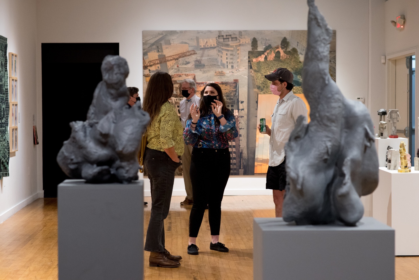 Two people speak in the gallery during the exhibition opening, with others viewing work; a pair of sculptures are in the foreground of the frame.